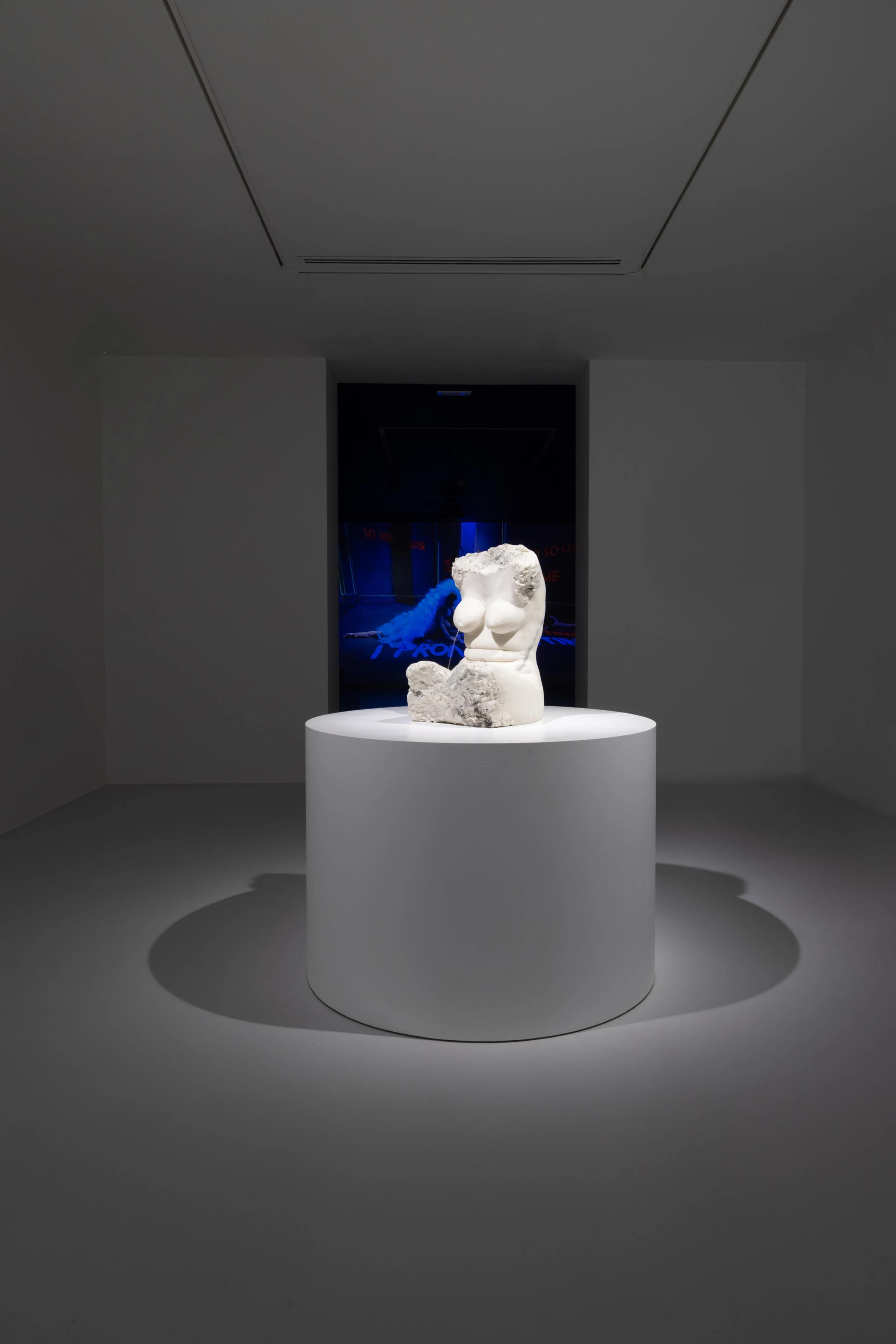 A pale sculpture on a circular plinth, with a blue projection behind it.